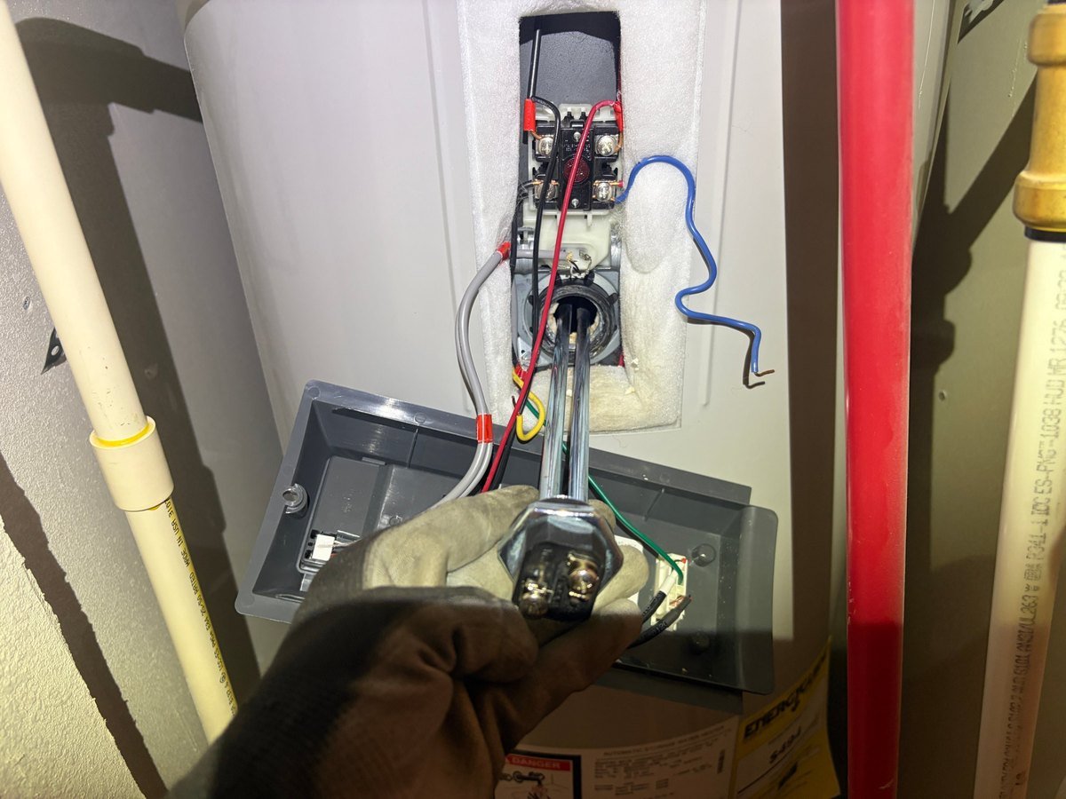 Gloved hand holding the removed lower heating element from a Bradford White electric water heater in Kennewick, with the opened access panel and its wiring behind it.